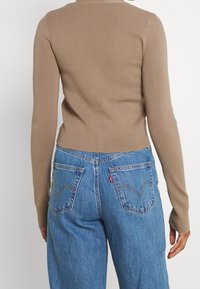 Person som bär en beige långärmad tröja och högmidjade blå jeans med bakfickor och en röd Levi’s etikett, sedd bakifrån.