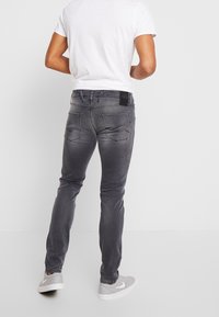 Pantalones vaqueros grises ajustados con diseño de cinco bolsillos. Textura desgastada, detalles de costuras visibles y una etiqueta con el logo en la cintura.