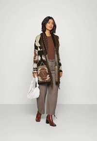 Žena nosi uzorčanu kardigan s resicama, smeđu rebrastu majicu, sive hlače, smeđe čizme s potpeticama do gležnja, drži bijelu torbu i stoji pred jednostavnom pozadinom.