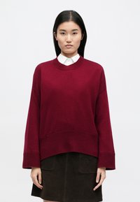Burgundský oversized svetr s žebrovaným výstřihem a manžetami, s texturovaným povrchem a uvolněným lemem, v kombinaci s tmavou sukní.