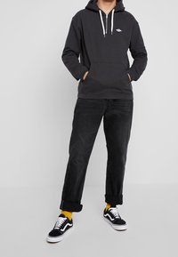 Musta huppari valkoisilla nyöreillä, etutasku, yhdistettynä tummiin farkkuihin ja mustiin lenkkikenkiin. Malli seisoo kädet taskuissa.