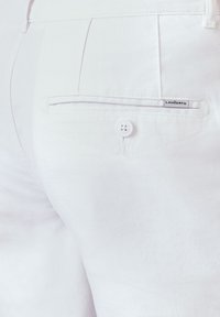 Białe bawełniane spodnie z tylną kieszenią zapinaną na guzik oraz małą metką z napisem "LANGERTO". Teksturowany materiał o delikatnym połysku.