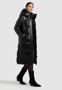 Abrigo tipo puffer negro con un cuello alto, diseño acolchado y bolsillos laterales, acompañado de botines de ante negros con punta afilada.