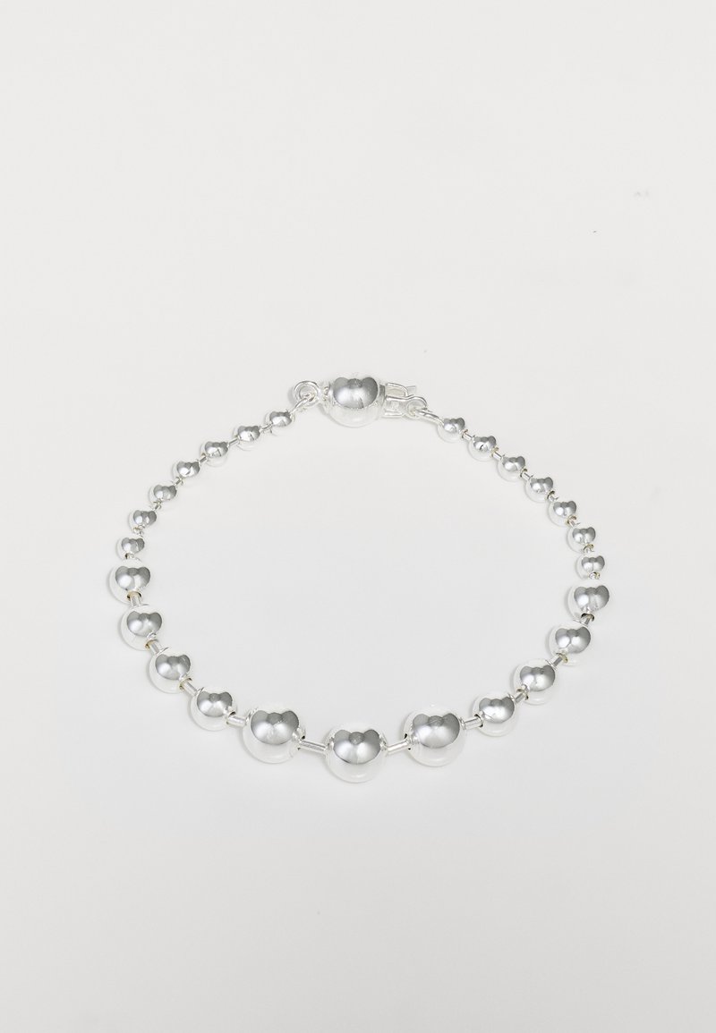 Sidabrinės spalvos apyrankė, kurią sudaro įvairaus dydžio sferiniai karoliukai, keičiantys mažomis metalinėmis jungtimis, užbaigta užsegimu. Lygi paviršiaus tekstūra.