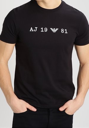 Man draagt een nauwsluitend zwart T-shirt met daarop in het wit "AJ 19" en "81", gescheiden door een wit gestileerd adelaarlogo op de borst.