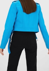 Zkrácená modrá umělá kožená bunda se strukturovanými švy a knoflíkovými detaily na rukávech, v kombinaci s černými přiléhavými kalhotami.