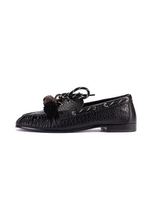 Mocasín de cuero negro texturizado con parte delantera cosida, lazo decorativo en negro y marrón, y tacón plano corto, mostrado en perfil lateral sobre fondo blanco.