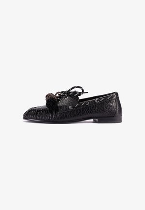 Mocasín de cuero negro texturizado con parte delantera cosida, lazo decorativo en negro y marrón, y tacón plano corto, mostrado en perfil lateral sobre fondo blanco.