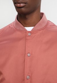 Close-up of person wearing a dusty pink buttoned jacket with ribbed collar over a white shirt, focusing on neck and upper chest.