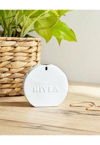 Weißer NIVEA-Kompaktcontainer auf einer Holzoberfläche neben goldenen Creolen und einem geflochtenen Pflanzenübertopf mit grünen Blättern.