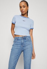 Lyseblå cropped t-skjorte med kontrastfarget blå krage og ermeender, med et lite grafisk motiv på brystet. Kombinert med høyt liv blå jeans.