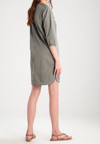 Harmaa puuvillamekko, jossa on kolmenneljänneksen hihat, pyöristetty helma ja sivuaukot. Mallilla on päällään litteät sandaalit. Neutraali väri ja yksinkertainen muotoilu.