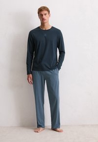 Man staat blootsvoets in een donkerblauw shirt met lange mouwen en losse blauwe pyjamabroek met patroon tegen een effen lichtgekleurde muur.