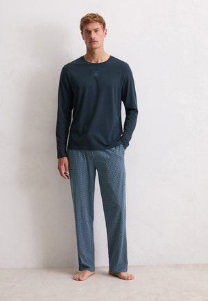 Man staat blootsvoets in een donkerblauw shirt met lange mouwen en losse blauwe pyjamabroek met patroon tegen een effen lichtgekleurde muur.