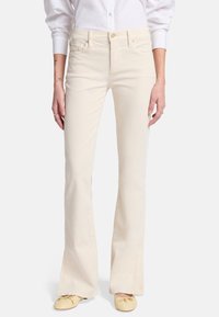 Witte flared jeans met een gladde textuur, vijf zakken, ritssluiting en goudkleurige hardware. Gecombineerd met lichtgele schoenen.