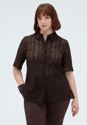 Vrouw met kort bruin haar die een donkerbruine blouse met patroon, korte mouwen en knopen draagt en bijpassende broek, staand met één hand in de zak.