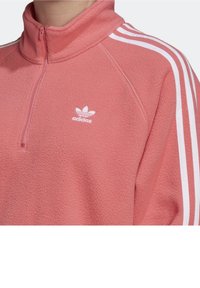 Pink fleece quarter-zip top with white three-stripe design on sleeves and small white Adidas logo on the chest. Soft texture, sporty fit.