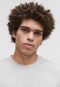 Svetlošedá košeľa s krátkym rukávom a zaobleným výstrihom, hladkou textúrou a jemným šitím na pleciach. Model má kučeravé tmavé vlasy.