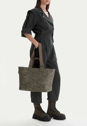 Mujer con mono negro y botas militares sosteniendo un gran bolso tote gris texturizado con asas dobles.