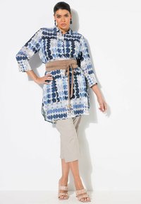 Knopfshirt-Tunika mit blau-weißem geometrischem Muster, kombiniert mit beigen Cropped-Hosen. Verfügt über einen gebundenen, beigen Stoffgürtel und bequeme beigen High Heels.