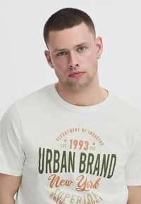 Krems krāsas īsām piedurknēm paredzēts T-krekls ar "URBAN BRAND NEW YORK" grafiku tumši zaļā un oranžā krāsā, apaļa kakla izgriezums, mīksts kokvilnas materiāls.