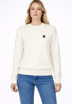 Blondynka w prostym kremowym longsleevie z małym czarno-złotym logo na piersi i niebieskich dżinsach.