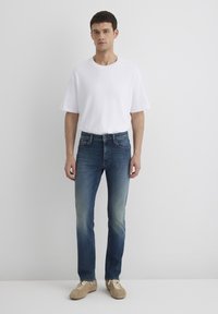 T-shirt bianca a maniche corte, jeans in denim blu con design sbiadito e sneakers beige. Il modello è in piedi frontale contro uno sfondo neutro.