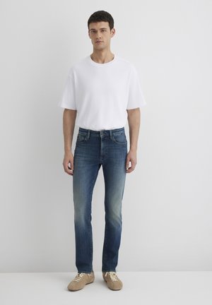 T-shirt bianca a maniche corte, jeans in denim blu con design sbiadito e sneakers beige. Il modello è in piedi frontale contro uno sfondo neutro.