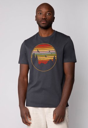 Donkergrijze t-shirt met korte mouwen, voorzien van een circulaire grafiek met horizontale strepen in oranje, geel en groen, en silhouetten van bomen en bergen.