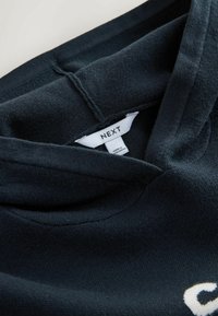 Čierna mikina s kapucňou vyrobená z jemného materiálu, s dizajnom v krku do V, ryhovanými detailmi a bielou nálepkou "NEXT" na vnútornej golierovej časti.