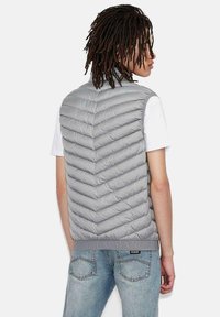 Grå pufferväst med ett chevron-mönster, hög krage och ribbat nederkant. Bärs över en vit skjorta med blå jeans.