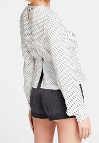 Blouse blanche à manches longues avec des pois noirs, présentant un détail en forme d'œillet dans le dos, un ourlet évasé, un tissu léger, portée avec un short noir.