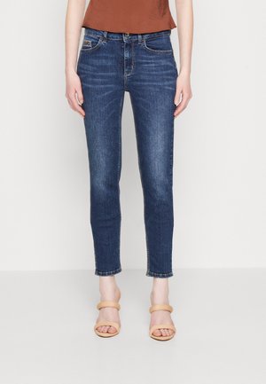 Personne portant un jean en denim bleu foncé ajusté et des sandales à talons beige, debout devant un fond clair uni.