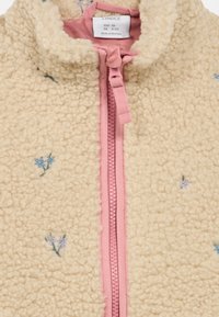 Veste en polaire de couleur crème avec une surface texturée. Dotée d'une fermeture éclair rose et d'une broderie florale bleue et verte sur le devant.