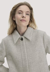 Hellgrauer Wollmischmantel mit weitem Kragen, der mit einem einzigen schwarzen Knopf geschlossen wird, und einem glatten, strukturierten Stoff.