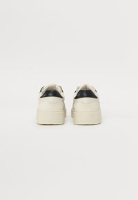 Baskets crème et noires avec une tige en cuir lisse, un col rembourré et une semelle en caoutchouc épaisse. Design minimaliste, montrant la vue arrière.