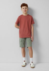 Rode katoenen T-shirt met korte mouwen, gecombineerd met groene cargoshorts met zijzakken. Witte sokken en beige sneakers maken de outfit compleet.