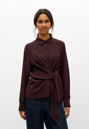 Une femme porte un chemisier boutonné bordeaux foncé, noué à la taille, et un jean foncé sur un fond blanc uni.