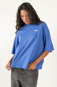 Blue cotton t-shirt with short sleeves and a crew neckline. Features a small white logo on the chest. Worn with dark gray jeans.