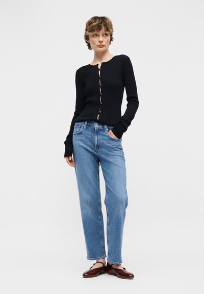 Γυναίκα με κοντά μαλλιά, που φοράει μαύρο πουκάμισο με κουμπιά και μακριά μανίκια, ανοιχτόχρωμο μπλε τζιν και σκούρα καφέ επίπεδα παπούτσια, στέκεται με το ένα χέρι στην τσέπη.