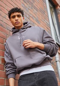Grå bomull hettegenser med frontlomme, snorhette og hvit logo. Brukt over en hvit skjorte med mørke denim jeans foran en murvegg.