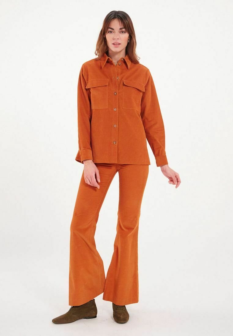 Chemise en velours côtelé orange et pantalon évasé assorti avec deux poches poitrine. Fermeture à trois boutons et col structuré. Bottes brunes à chevilles.