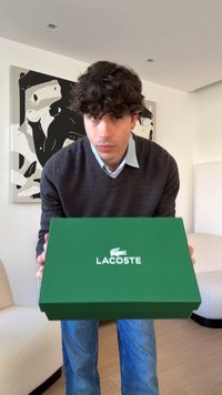 Homme aux cheveux bouclés foncés portant un pull gris et un jean bleu tenant une boîte à chaussures verte Lacoste vers la caméra dans un salon moderne.