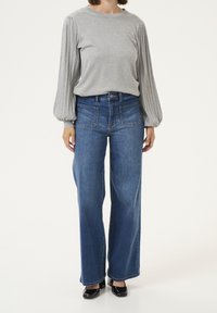 Pull-over gris avec manches ballon côtelées, associé à un jean large bleu taille haute. Des chaussures à talons foncés complètent la tenue.
