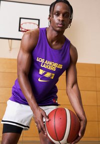 Lila kosárlabda mez, amelyen a "LOS ANGELES LAKERS" felirat sárgával szerepel, fehér sorttal párosítva. A személy egy kosárlabdát tart egy edzőterem környezetében.
