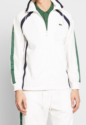 Mann trägt weiße Trainingsjacke mit Reißverschluss und Hose mit dunkelgrünen und schwarzen Streifen, darunter grünes Poloshirt.