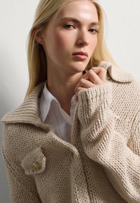 Beige knitted cardigan featuring a wide collar, ribbed texture, and a gold button accent on the front. Worn over a white collared shirt.