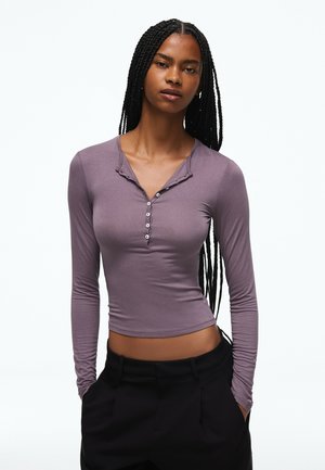 Jeune femme avec de longues tresses portant un haut ajusté lavande à manches longues boutonné et un pantalon noir, les mains dans les poches.