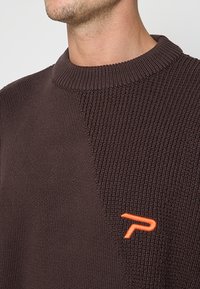 Pull en tricot marron avec un motif texturé, doté d'un col rond et d'un accent logo orange sur le côté gauche.