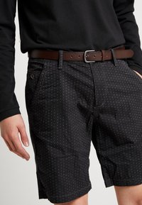 Pantalones cortos negros con un patrón texturizado de pequeños puntos blancos, con un cinturón de cuero marrón y bolsillos laterales. Material mezcla de algodón.